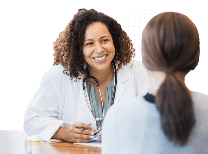 A smiling female doctor wearing a white coat and stethoscope is sitting at a desk and conversing warmly with a patient
